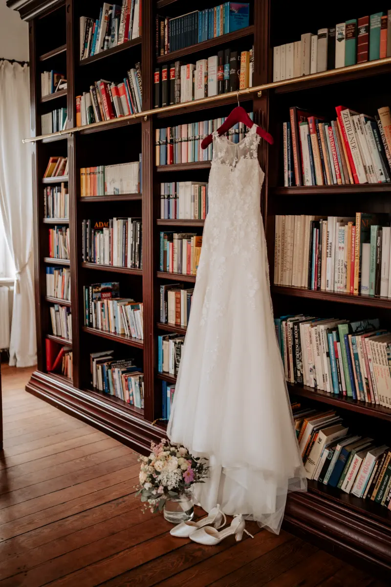 Ein Brautkleid, aufgehängt vor einer Bücherwand im Salon, mit Brautstrauß und Schuhen.