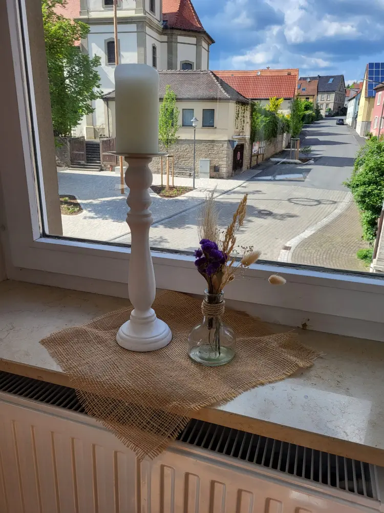 Minimalistische Fensterdekoration mit einer weißen Kerze und einer Blumenvase auf einem rustikalen Untersetzer im Salon.