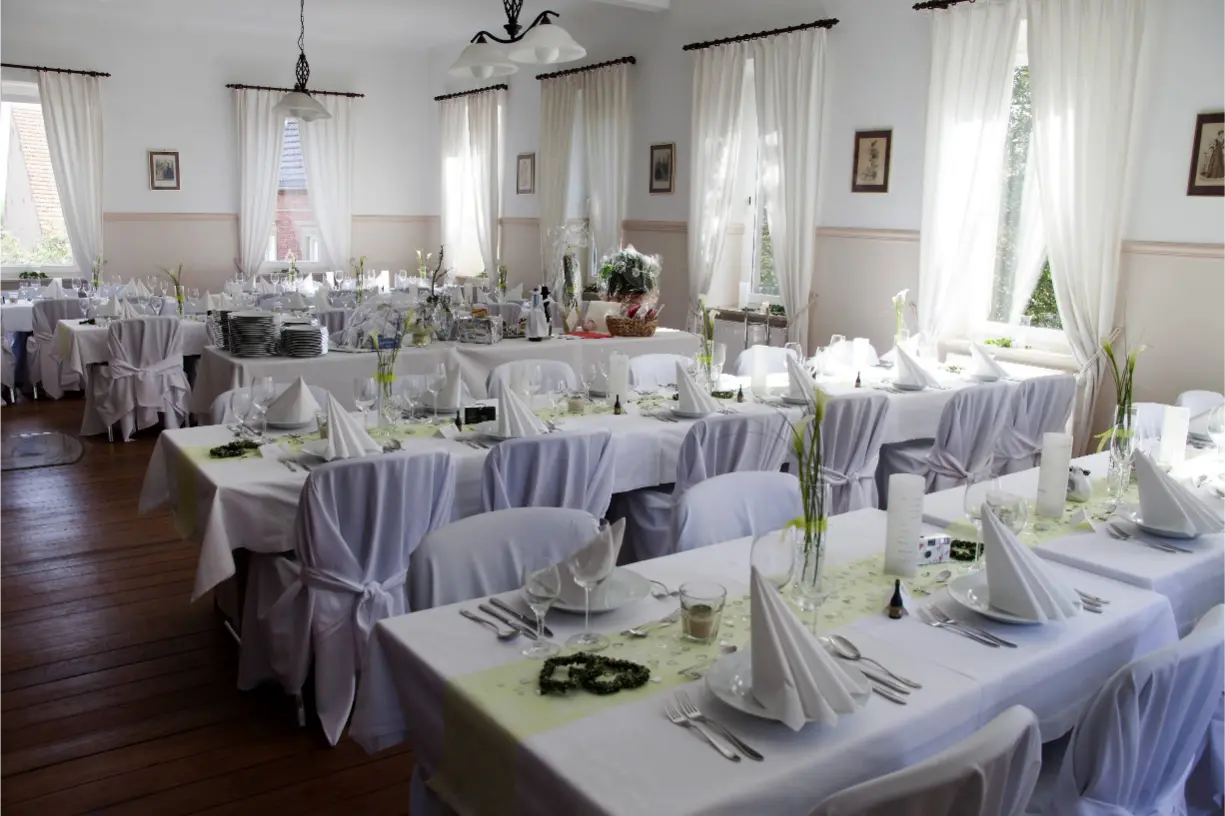 Eleganter Hochzeitssalon mit liebevoll eingedeckten Tischen, weißen Stuhlhussen und natürlicher Dekoration.